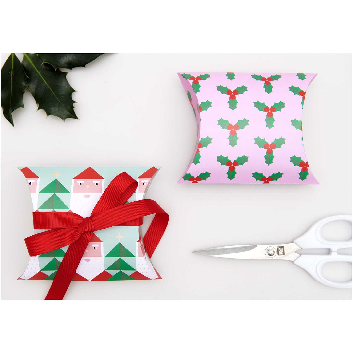 Paper Poetry Geschenkschachteln Merry Christmas groß mit festlichen Motiven und rotem Band, dekoriert neben einer Schere und Stechpalmenblättern.