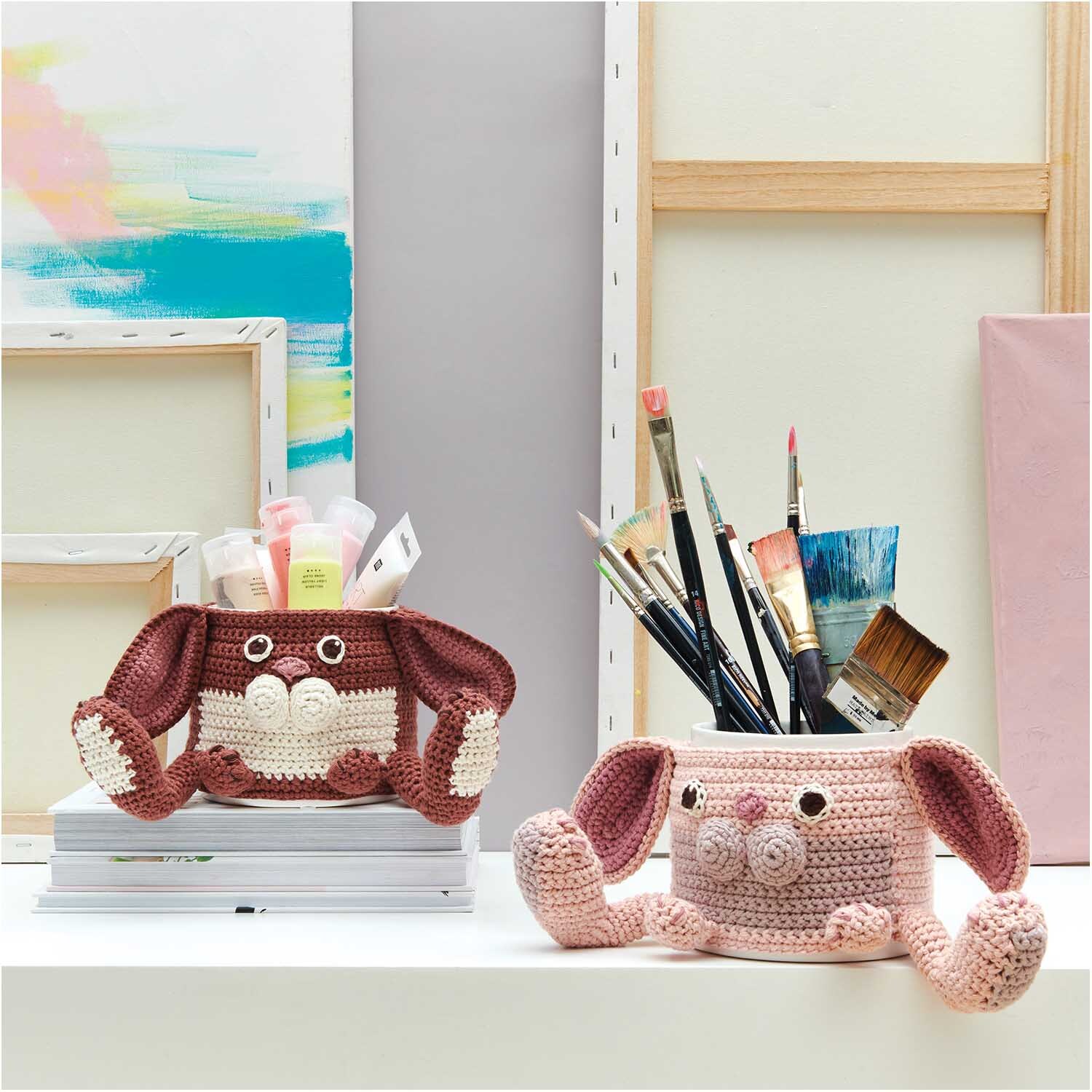 Creative Cotton aran: Zwei gehäkelte Stifthalter in Tierform, gefüllt mit Pinseln und Stiften, auf einem Tisch.