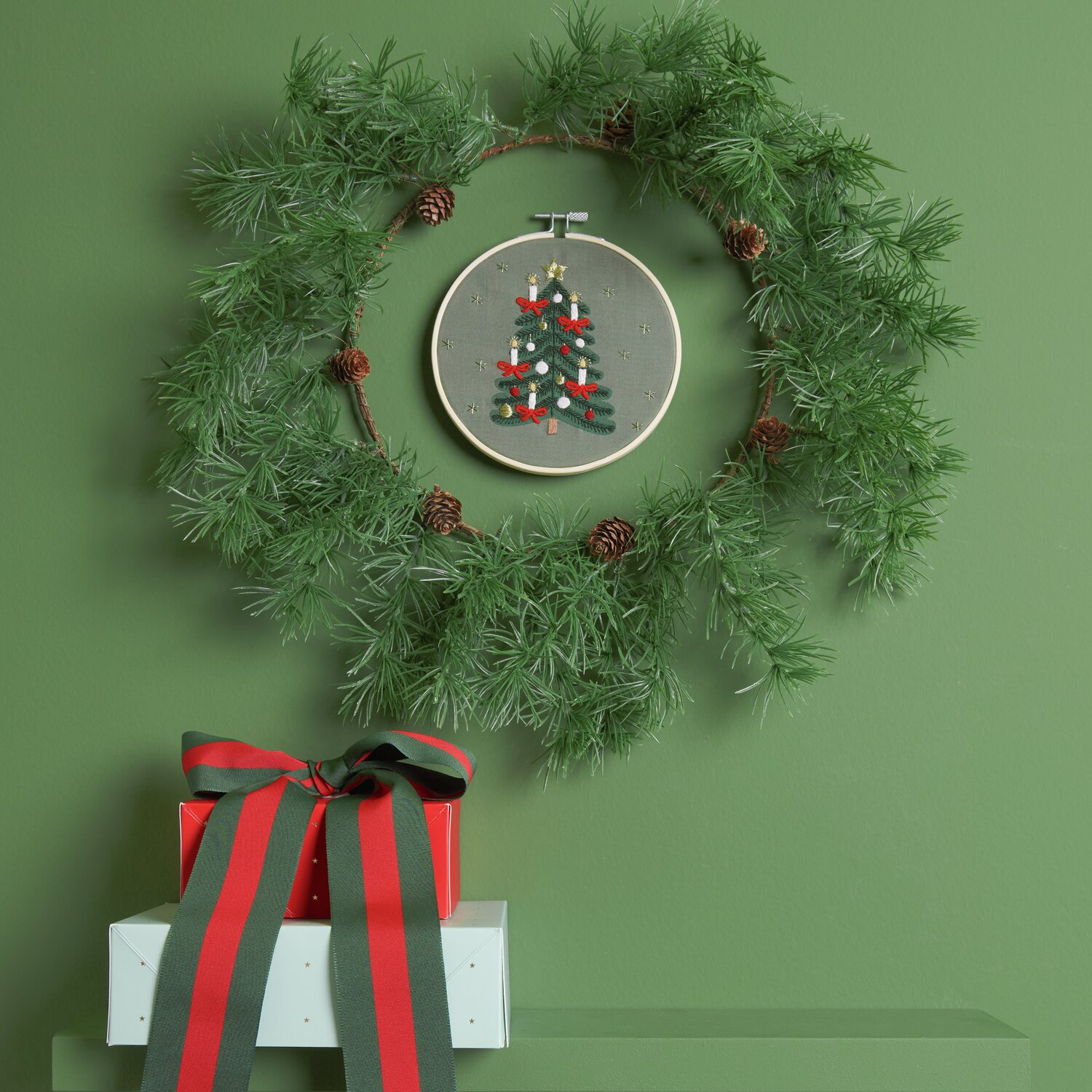 Stickpackung Weihnachtsbaum vorgezeichnet: Ein grüner Kranz mit Stickrahmen und Weihnachtsbaum-Motiv, dekoriert mit Geschenken.