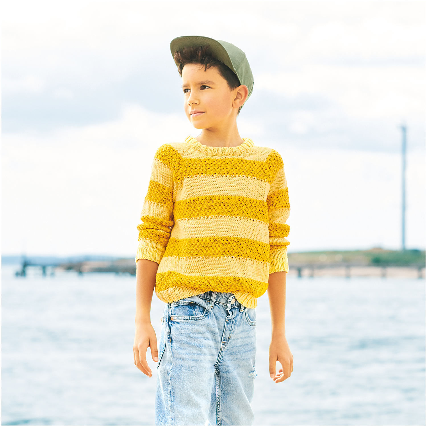 Essentials Organic Cotton aran: Junge am Strand in gelbem Strickpullover von Rico Design, kombiniert mit Jeans und Mütze.