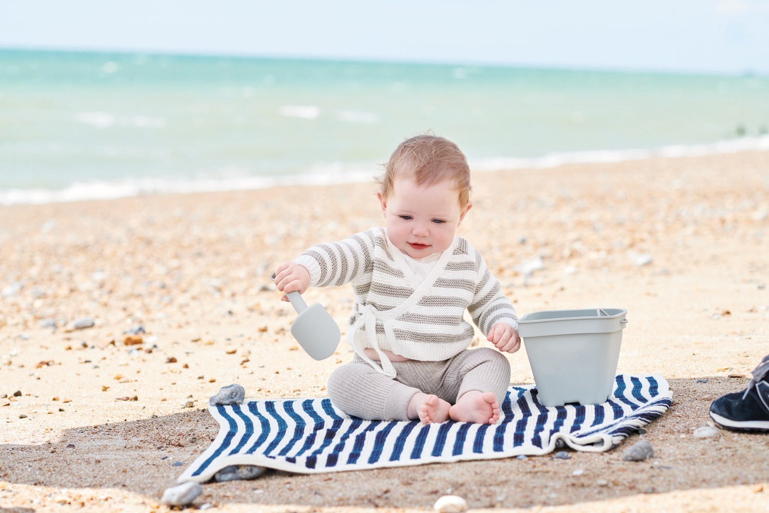 Baby Organic Cotton Strickset Decke am Strand mit Baby in gestreiftem Outfit und grauem Eimer.