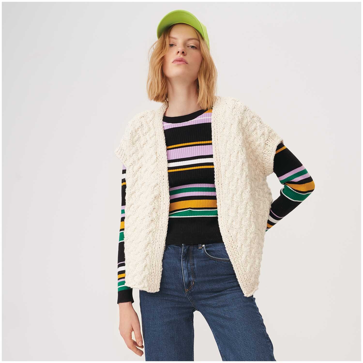 Creative So Cool + So Soft Cotton chunky: Frau trägt cremefarbene Strickweste von Rico Design über gestreiftem Pullover und Jeans, mit grünem Cap.