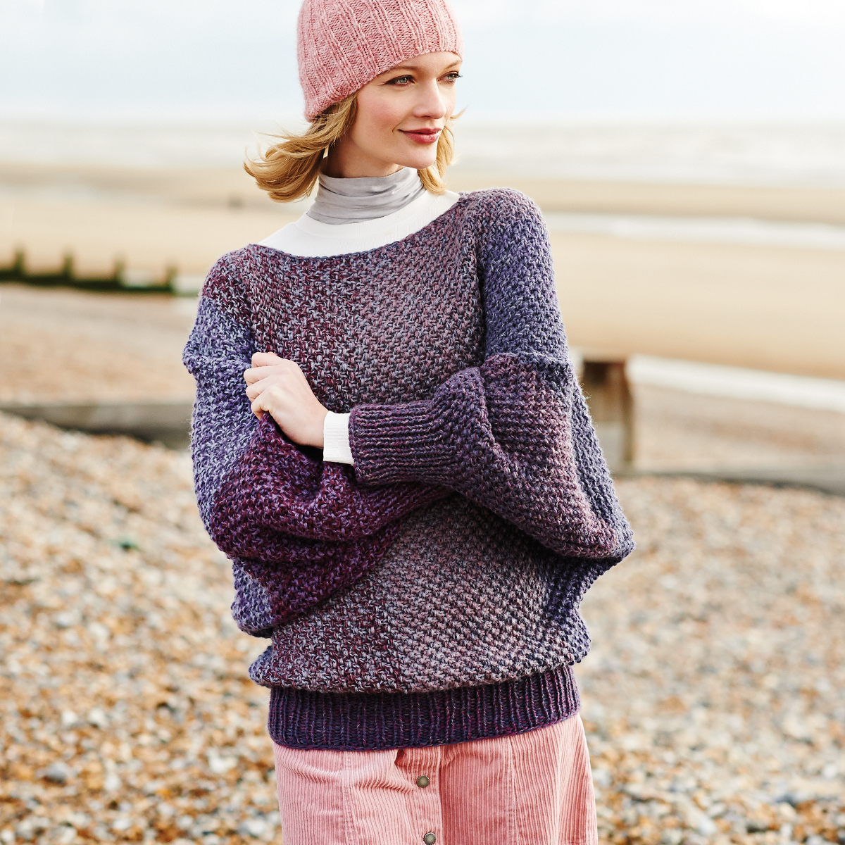 Strickanleitung Pullover mit Perlenmuster: Frau am Strand trägt lila Pullover und rosa Mütze.