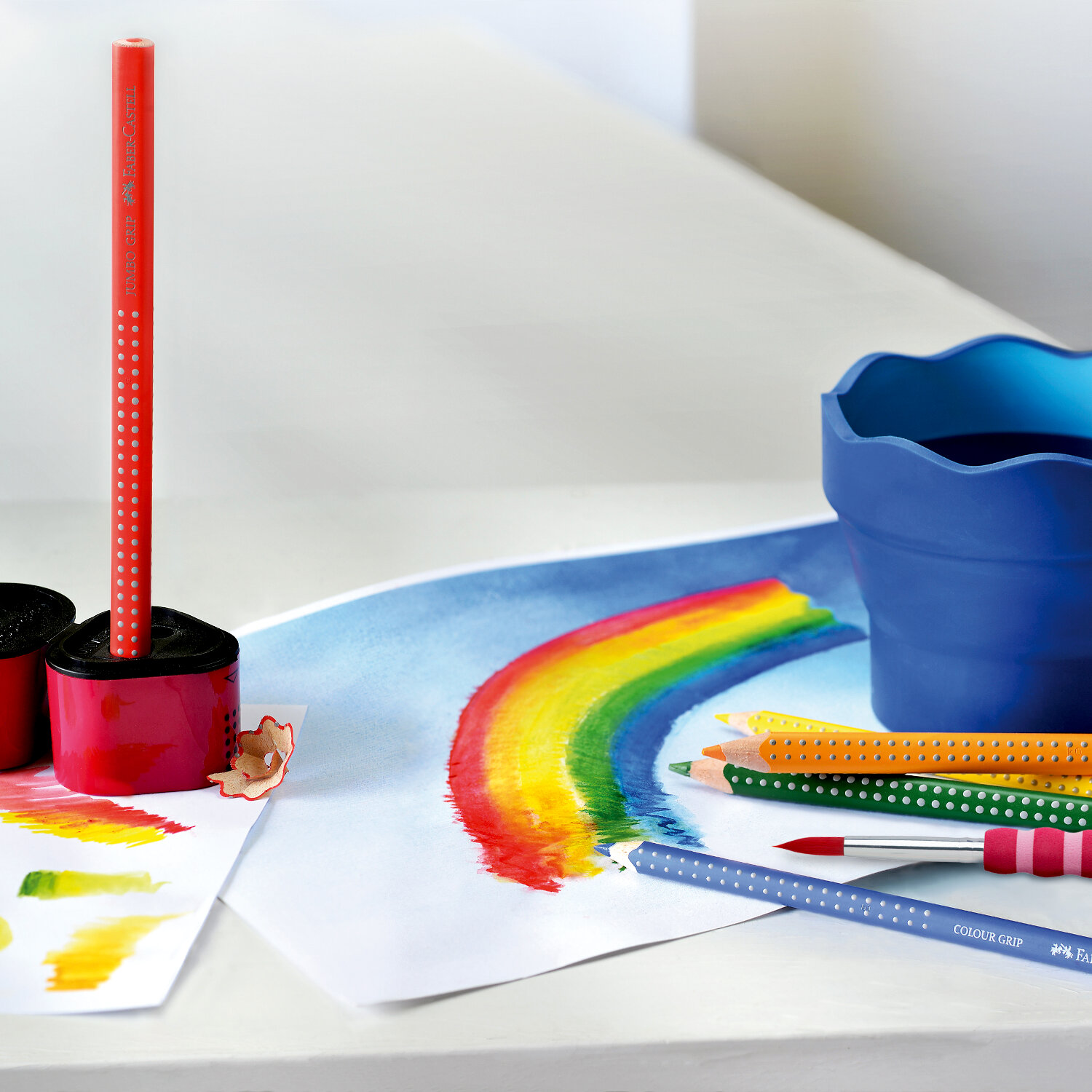 Colour Grip 2001 Buntstifte von Faber Castell auf einem Tisch mit Regenbogenzeichnung und Farbtöpfen.