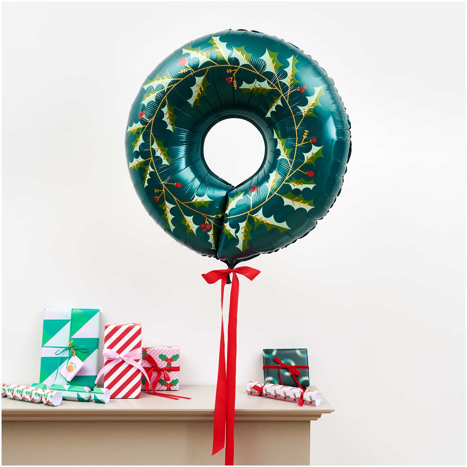 Folienballon Weihnachten Kranz 71x20cm in Grün mit Stechpalmenmotiv, dekoriert mit roten Bändern, schwebt über Geschenken auf einem Tisch.