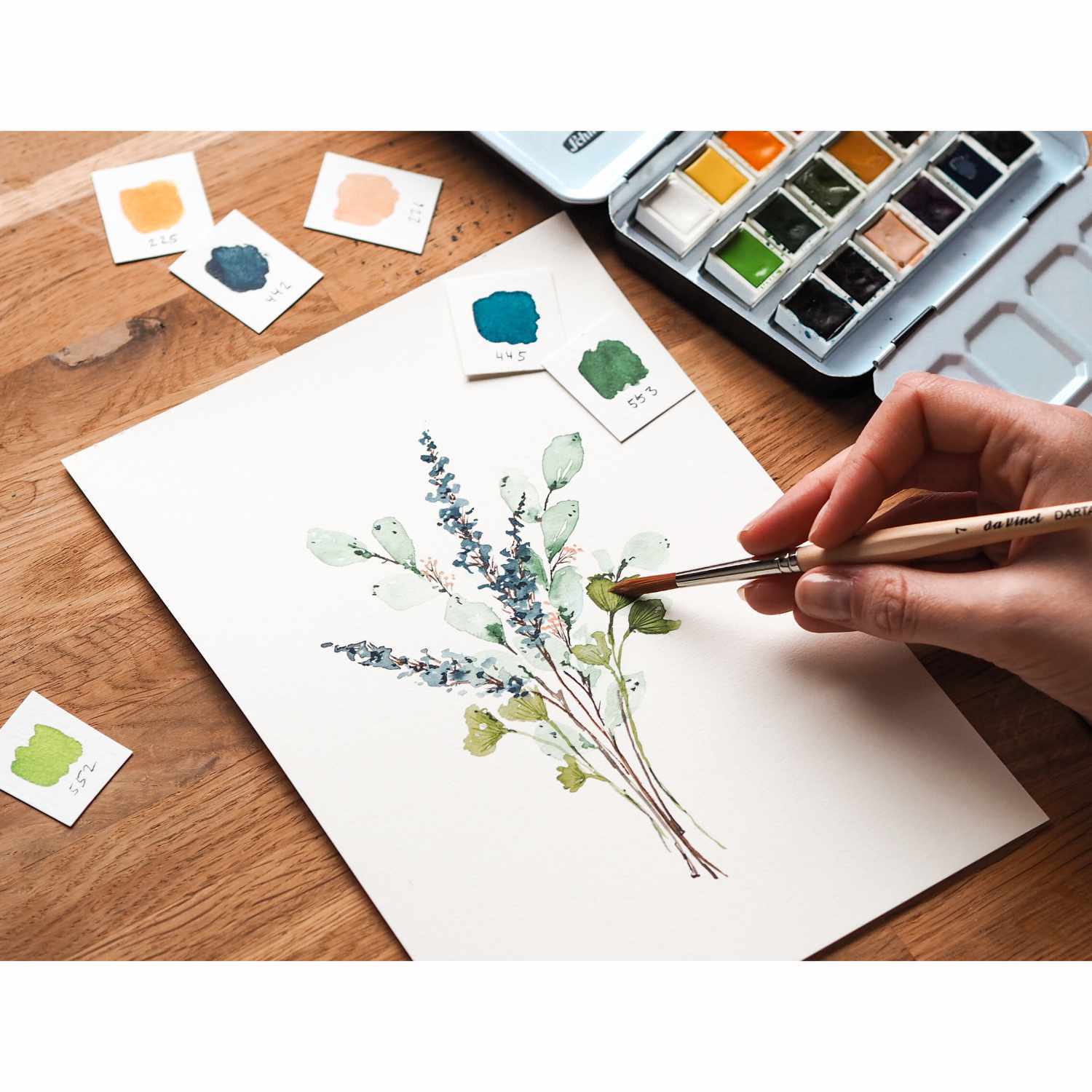Schmincke Akademie Aquarellkasten mit 18 Farben. Eine Hand malt Pflanzen auf Papier, umgeben von Farbmusterkarten.
