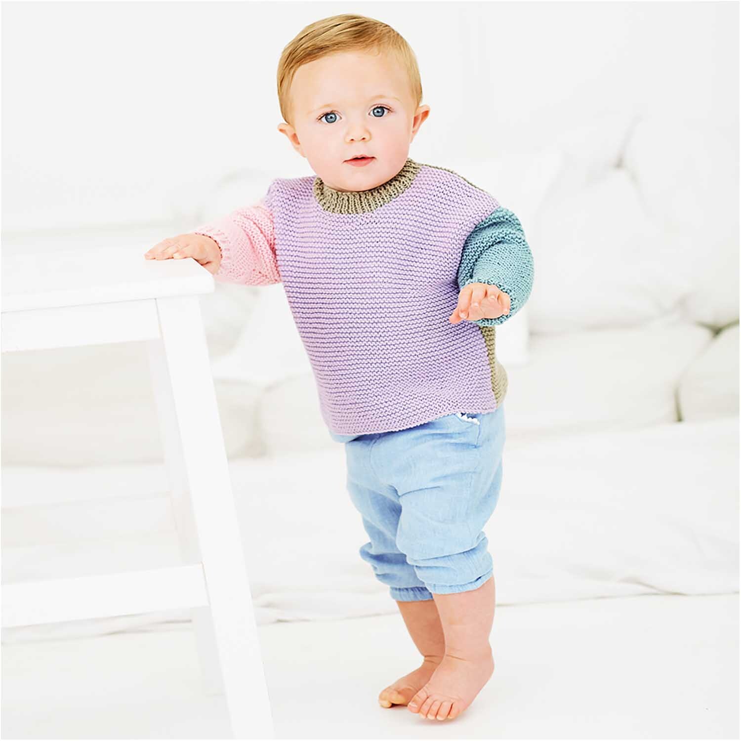 Rico Baby Cotton Soft dk: Ein Baby trägt einen gestrickten Pullover in Pastellfarben mit blauen Hosen.