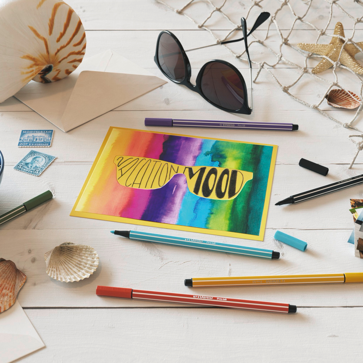 Pen 68 von STABILO liegt auf einem Tisch mit buntem Kunstwerk, Muscheln und Sonnenbrille.