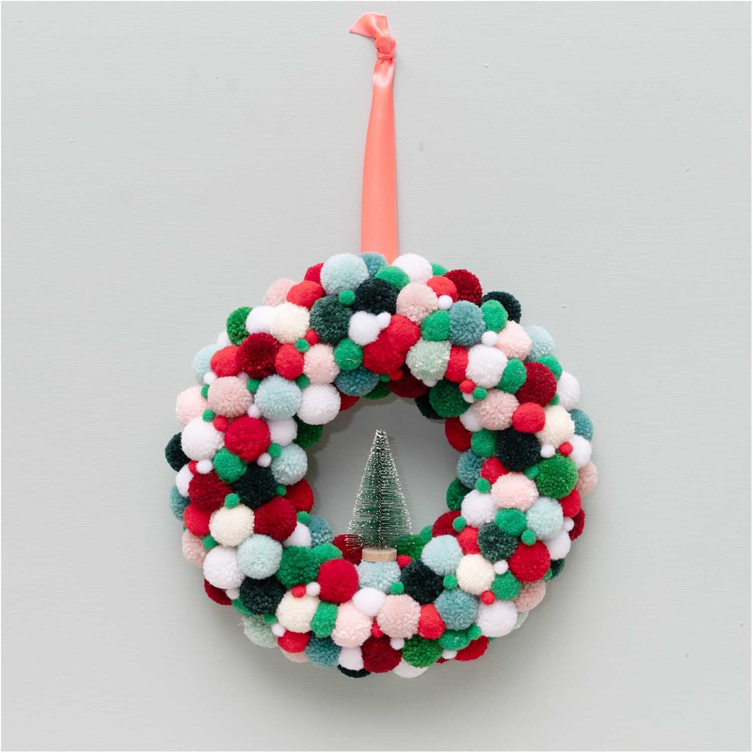 Garn Pompons 24 Stück: Kranz aus bunten Pompons mit Rot-Grün Mix, aufgehängt an einem rosa Band.