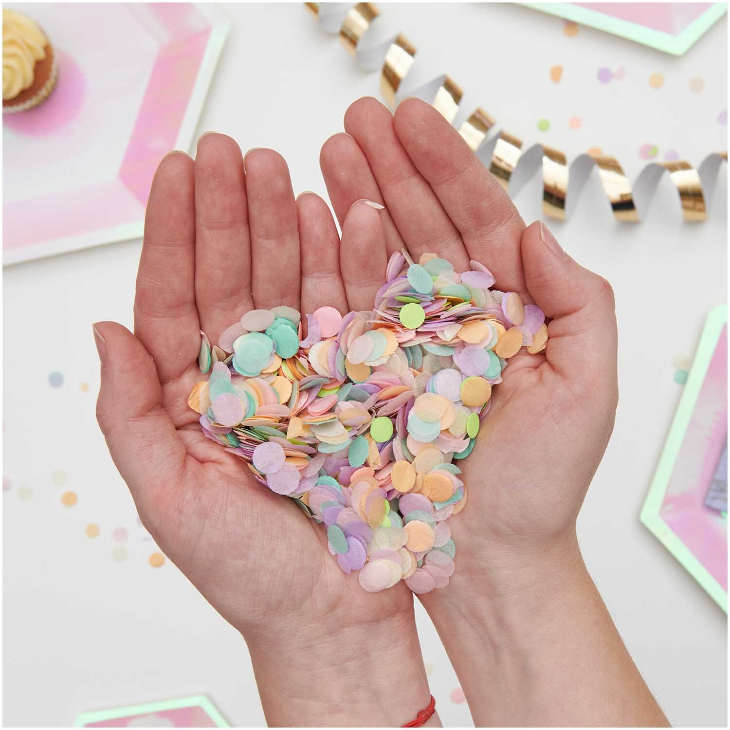 Konfetti Pastell Rainbow Mix 20g in Händen gehalten, umgeben von Partydekorationen, ideal für Feiern.