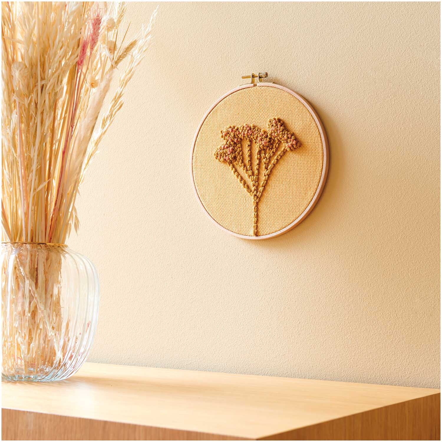 Punch Needle Packung Bild Schleierkraut zeigt ein gesticktes Blumenmotiv in einem Holzrahmen an einer beigen Wand. Links steht eine Vase mit Trockenblumen.