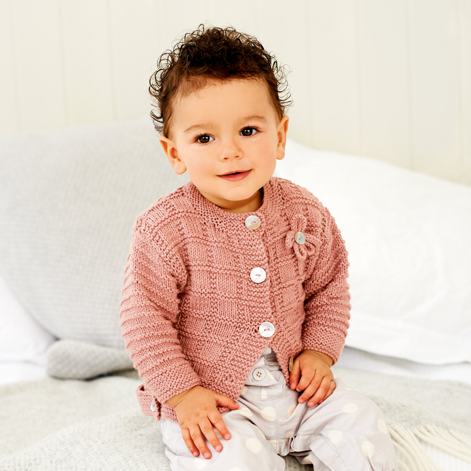 Strickanleitung Babyjacke mit Rundhalsausschnitt, detailreich gestrickte roséfarbene Babyjacke mit weißen Knöpfen, getragen von lächelndem Kleinkind auf Bett
