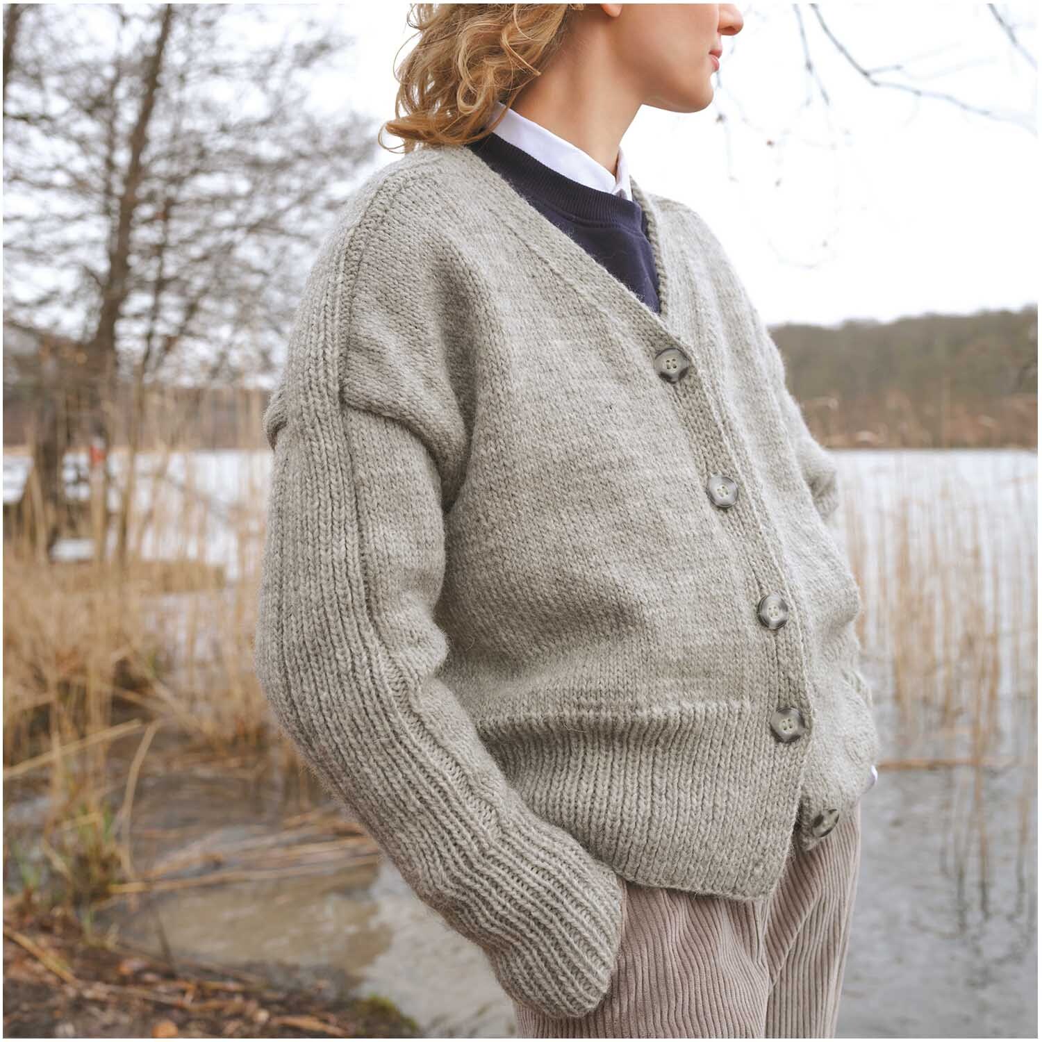 Essentials Organic Wool aran: Person trägt einen gestrickten Cardigan am Seeufer, umgeben von Schilf.