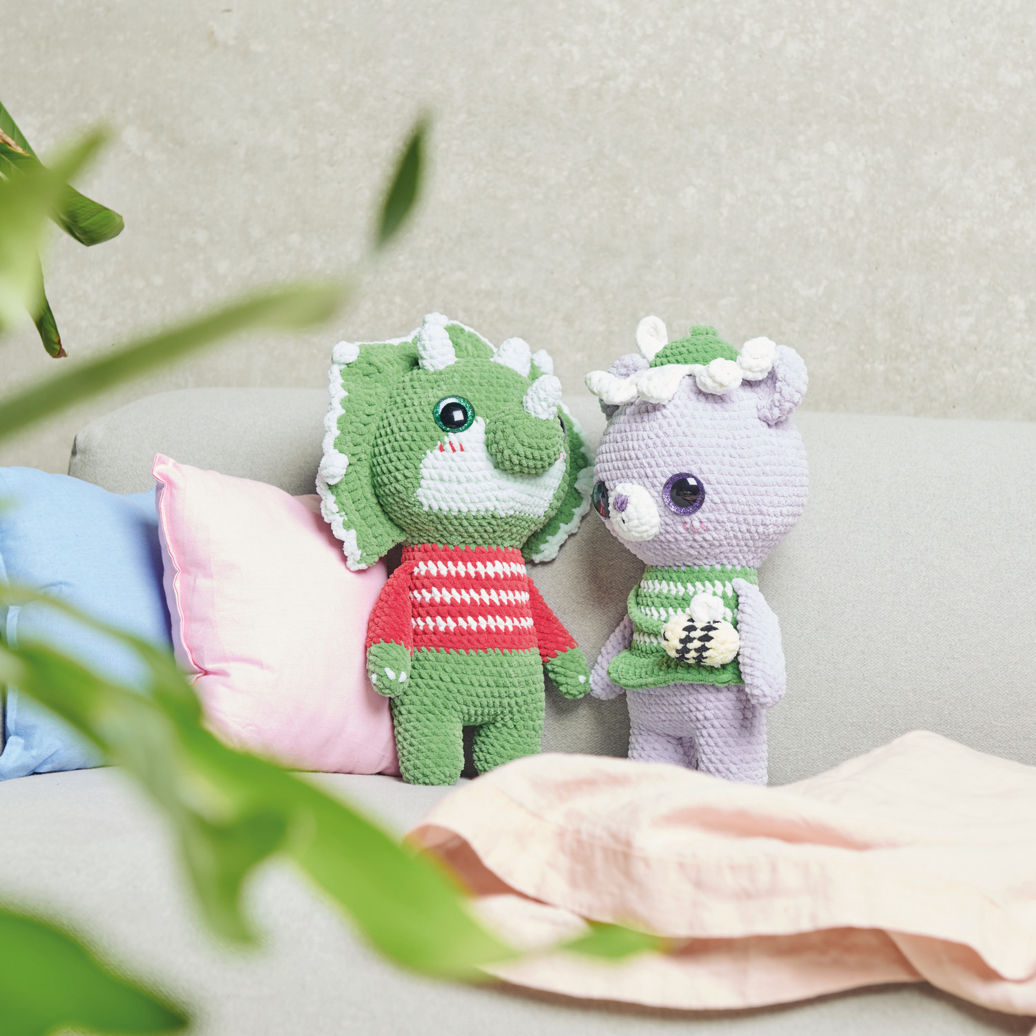 Häkelset Baby Dinosaur Modell 01 und Flower Lover Bear Modell 07 aus Chenillove Big Cuties auf einem Sofa mit bunten Kissen.