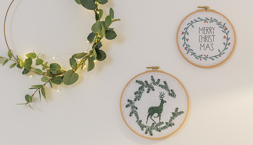 Stickanleitung Weihnachtliche Homedeko im Stickring: Zwei bestickte Ringe mit Rentier und Weihnachtsgruß, umgeben von grünen Zweigen.