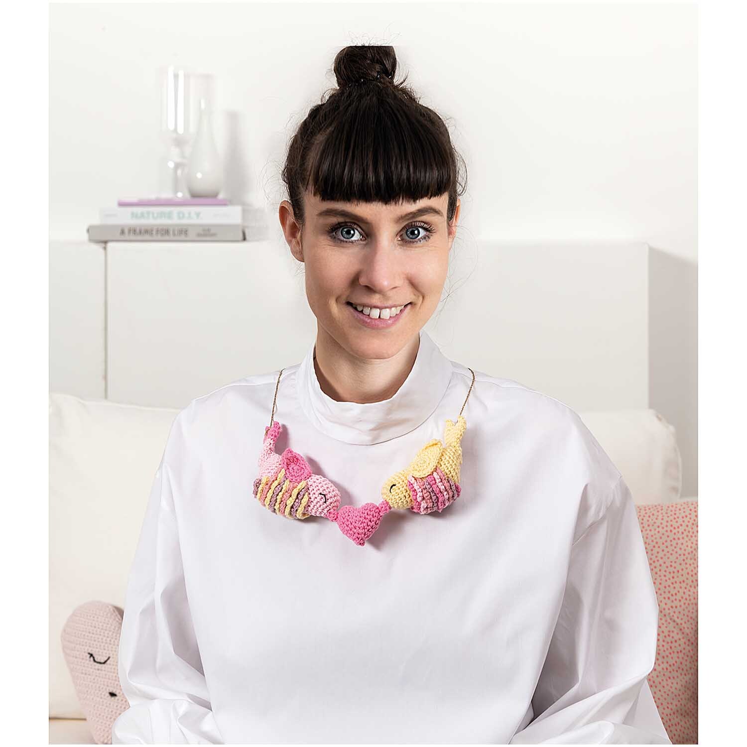 Creative Ricorumi dk: Frau mit gestrickter Halskette in Pastelltönen, lächelnd in modernem Raum.