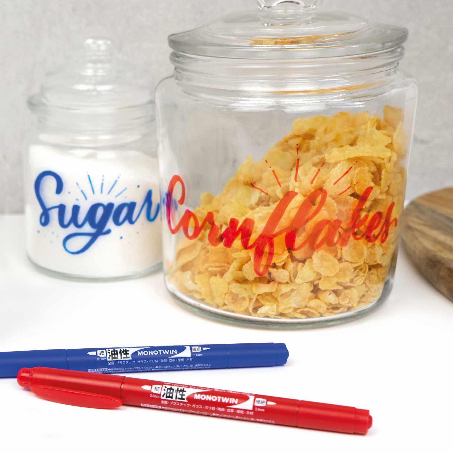 Fineliner MONO twin: Zwei Stifte in Blau und Rot vor beschrifteten Vorratsgläsern mit Zucker und Cornflakes.