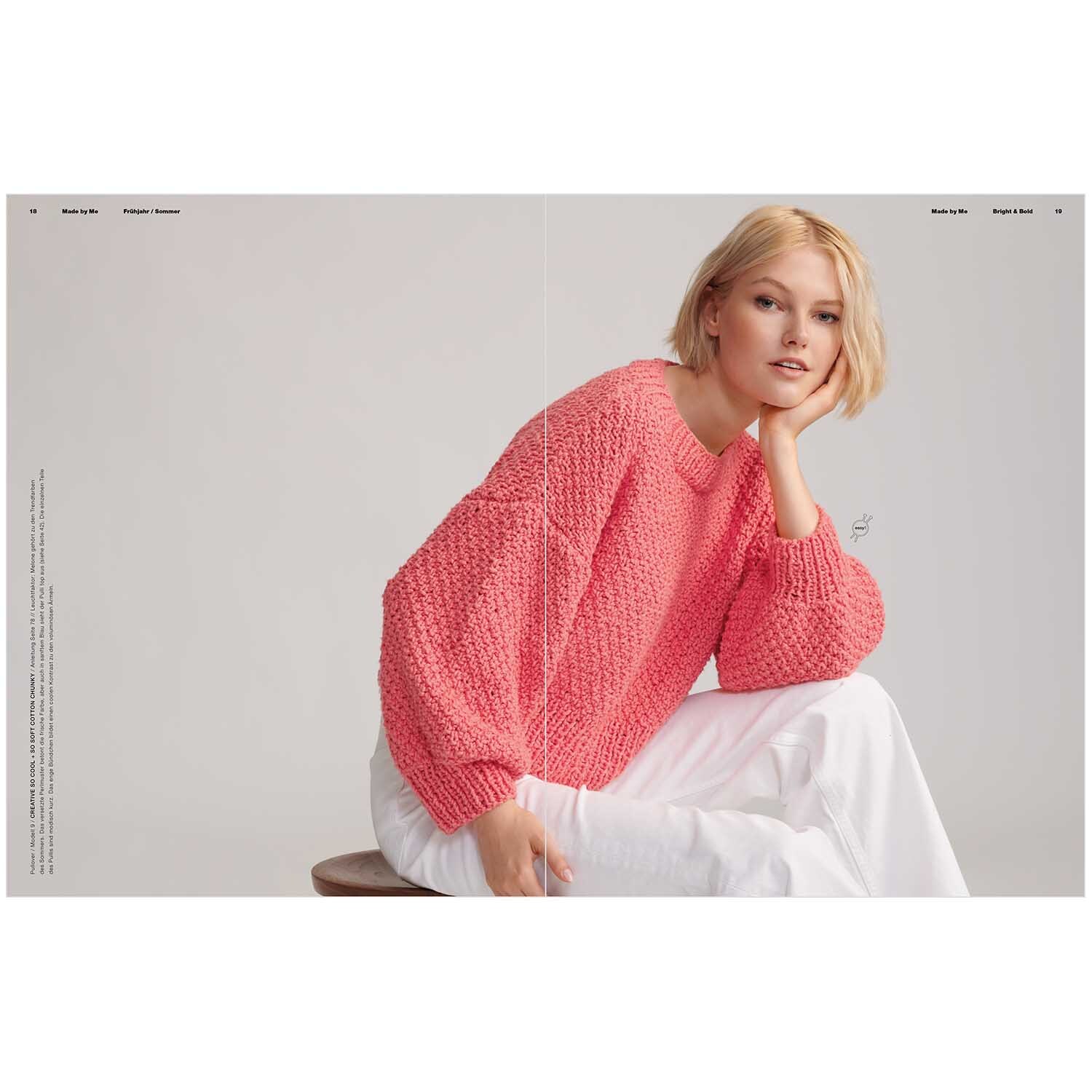 Made by Me Handknitting Nr. 12 Frühjahr-Sommer: Frau in rosa Strickpullover von Rico Design, sitzt auf Hocker vor grauem Hintergrund.