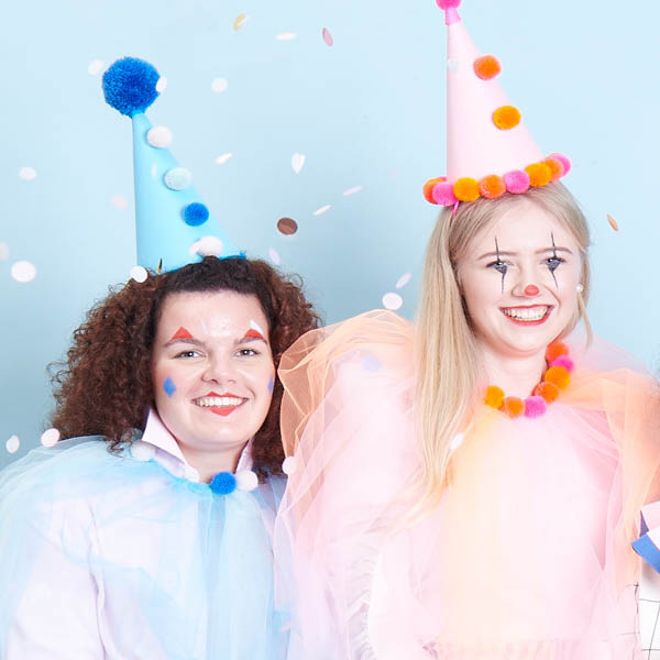 Zwei Frauen, kostümiert als blauer und pinker Clown, lächeln. Fotosetting blaue Wand und Konfetti.
