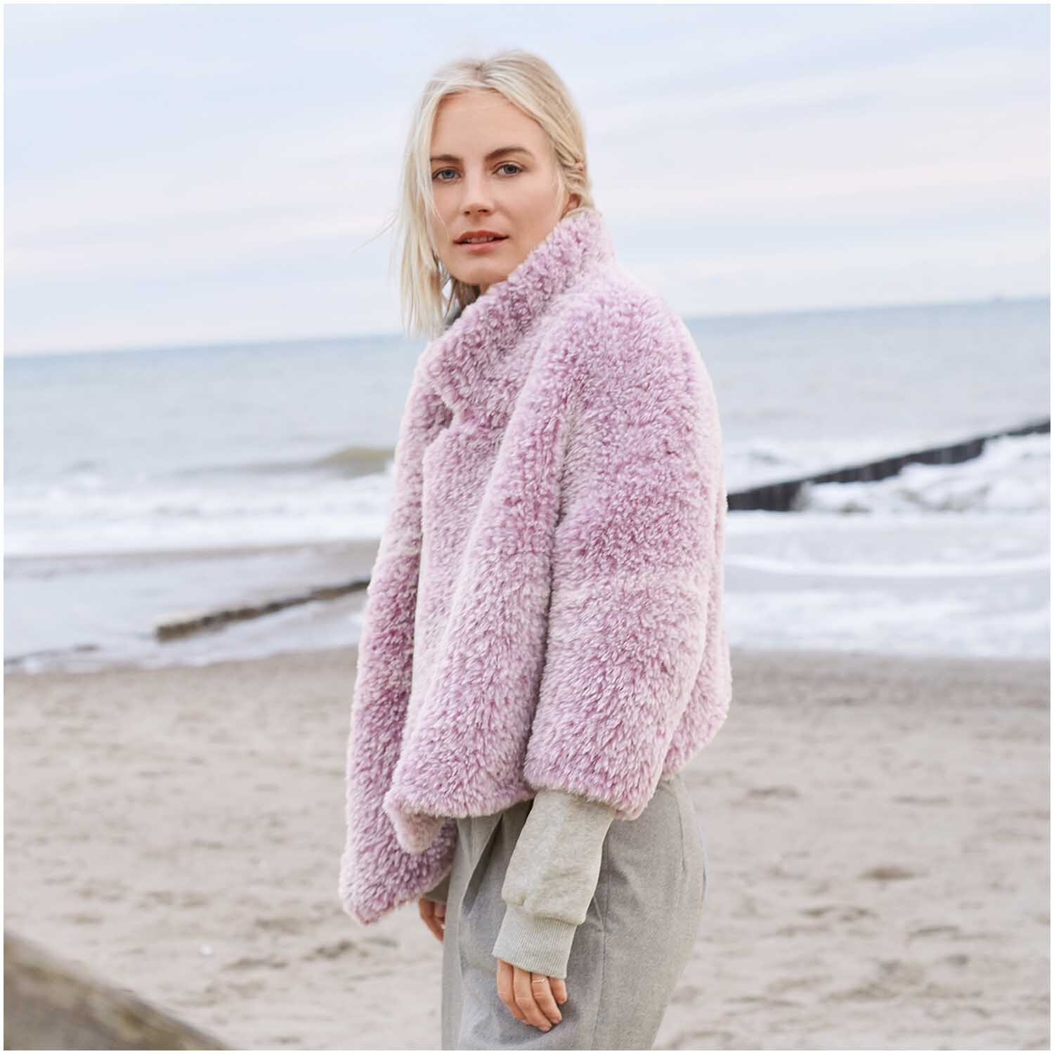 Creative Petit Lapin Super Chunky: Frau am Strand in fliederfarbener Jacke von Rico Design, umgeben von sanften Wellen.