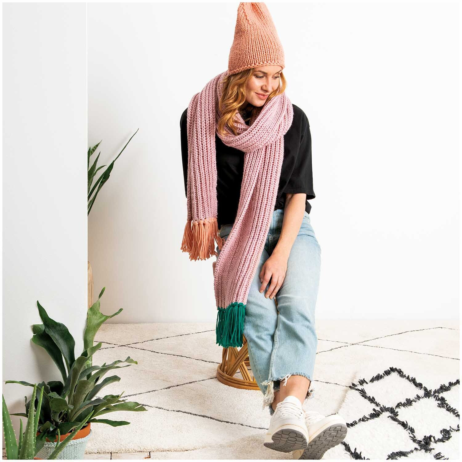 Essentials Mega Wool chunky: Frau trägt rosa Schal und Mütze, sitzt auf Hocker, umgeben von Pflanzen.