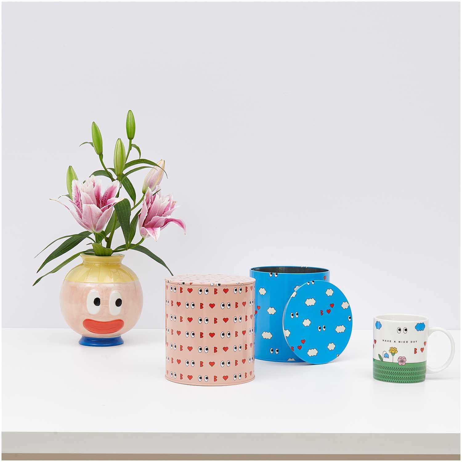 Rico Design x Redfries Keksdose Liebe rosa 13,5x15cm auf einem Tisch mit Blumen in einer Vase, einer blauen Dose und einer Tasse.