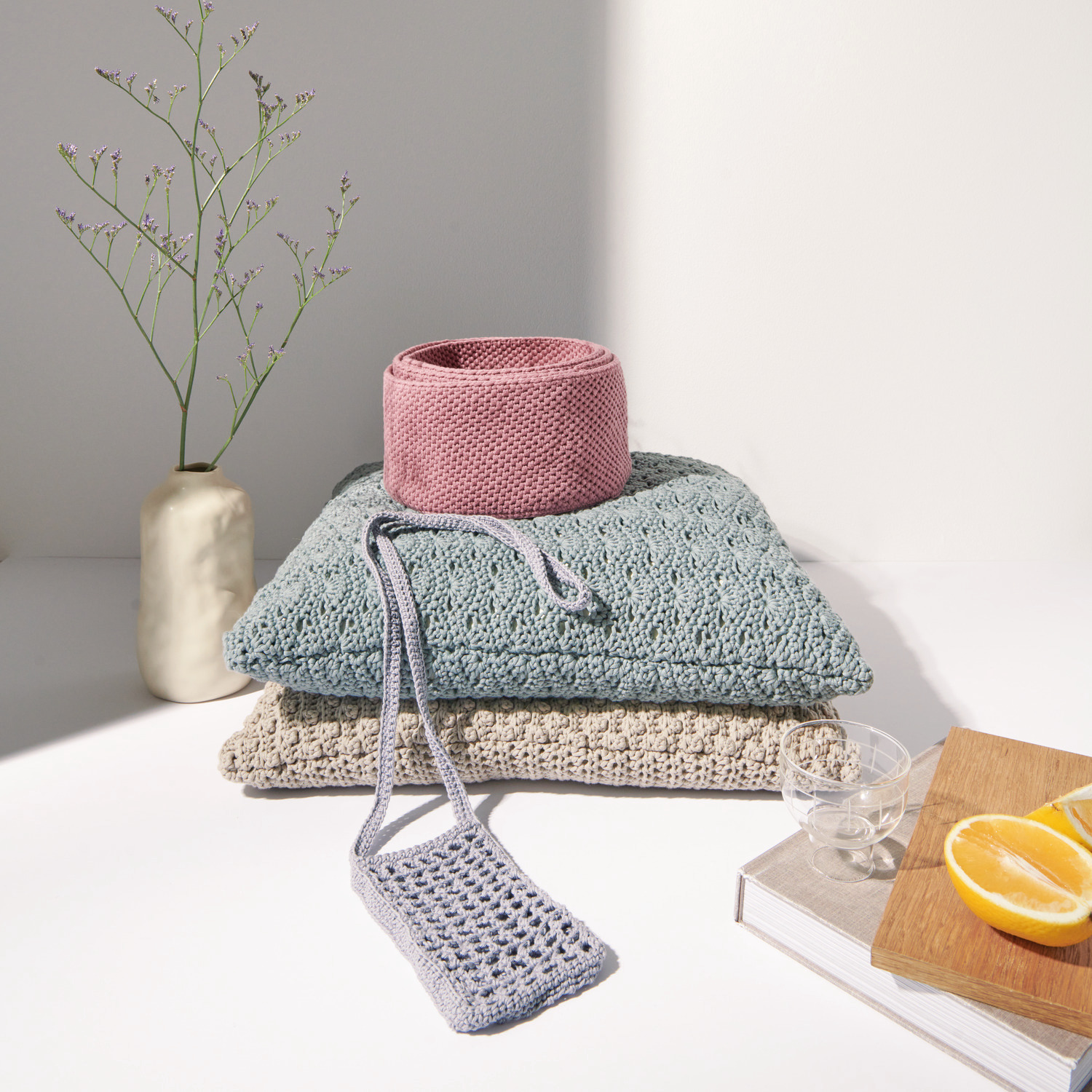 Häkelset Kissen Modell 08 aus Boho Crochet: Drei gehäkelte Kissen in sanften Farben, dekoriert mit einem rosa Korb und einer grauen Handytasche.