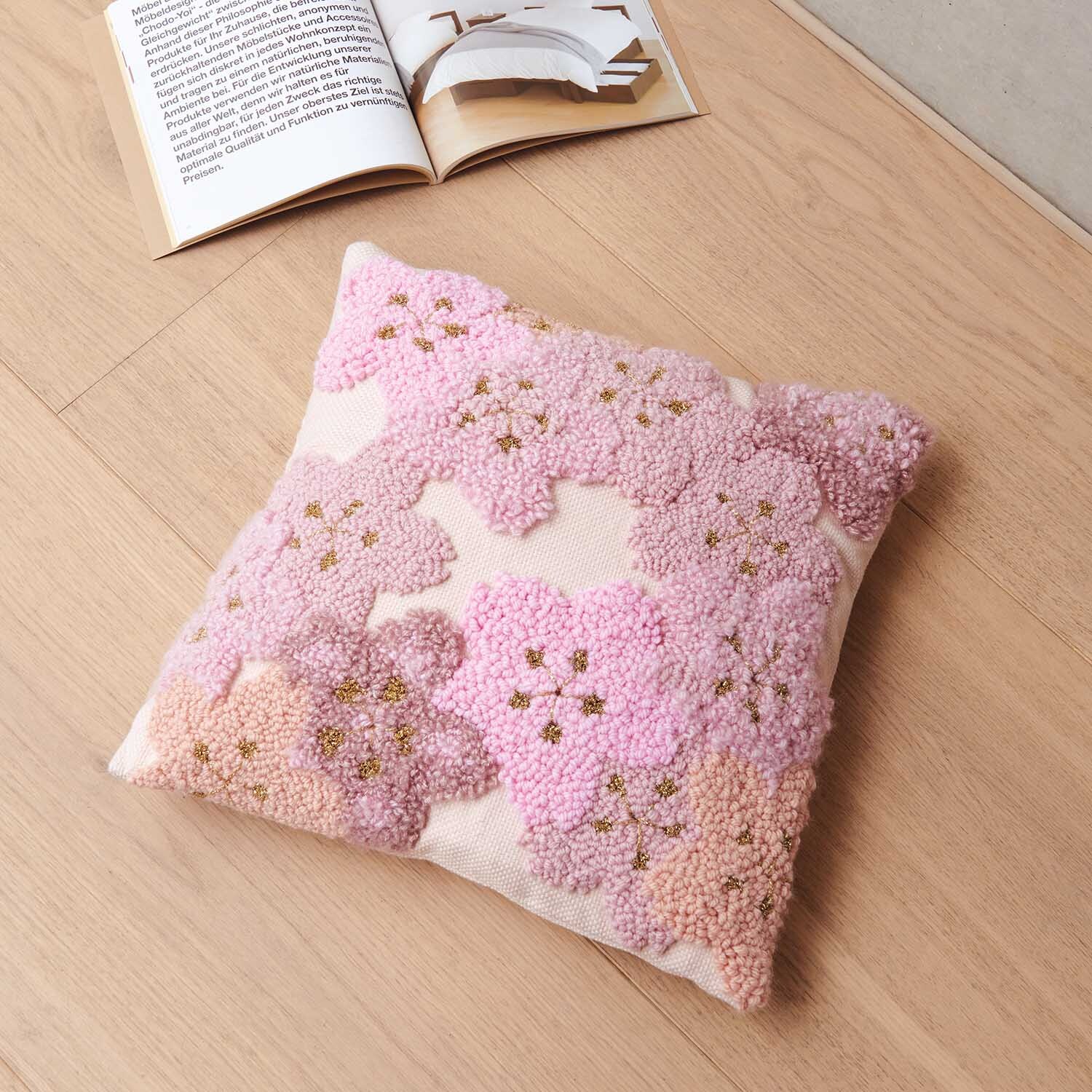 Anleitung Kissen mit Kirschblüten punchen: Ein Kissen mit rosa Kirschblütenmuster auf Holzfußboden neben einem offenen Buch.