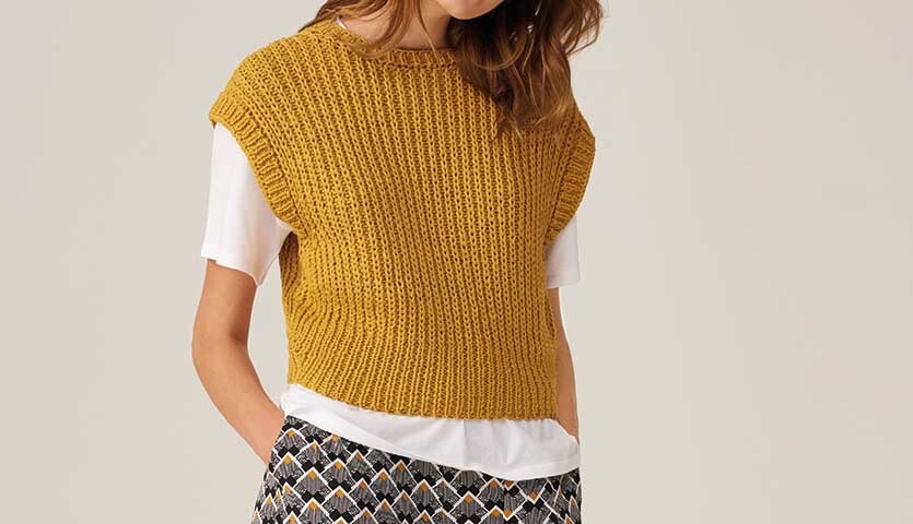 Strickanleitung Top aus Fashion Cotton Ribbon chunky: Frau trägt gelbes Stricktop über weißem Shirt, kombiniert mit gemusterten Hosen.