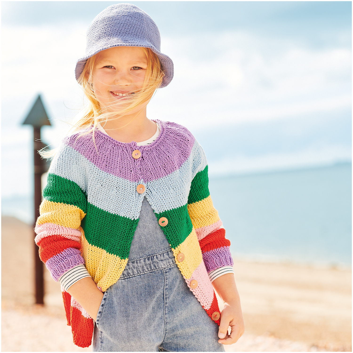 Essentials Organic Cotton aran: Kind in bunter Strickjacke am Strand, trägt lila Hut, lächelt fröhlich.