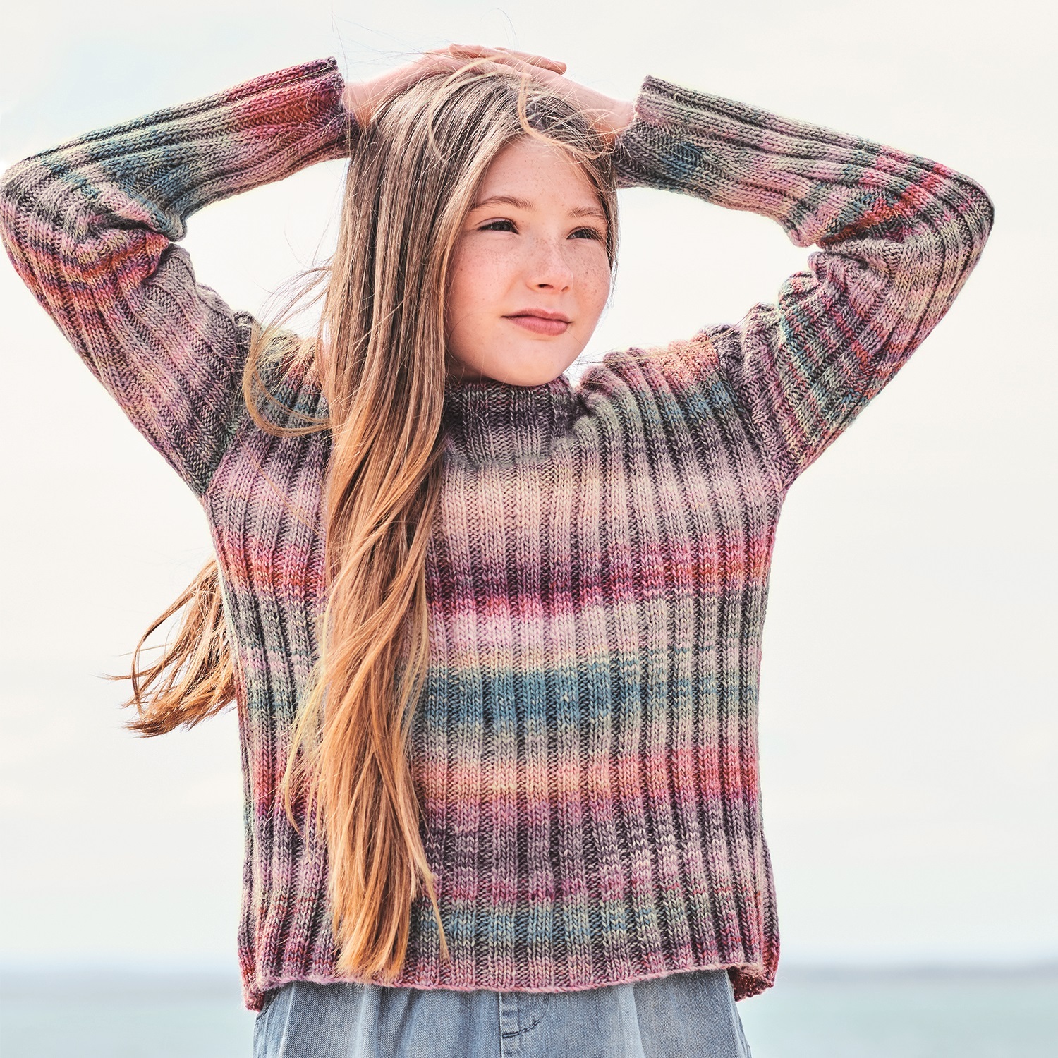 Strickanleitung Kinderpullover mit Farbverlauf: Mädchen trägt gestreiften Pullover in sanften Tönen am Strand.