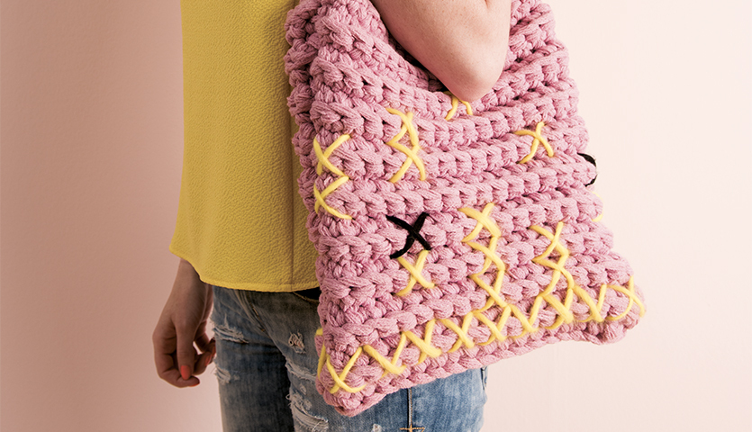 Häkelanleitung bestickte Tasche aus Creative Loopy: Rosa Tasche mit gelben Stickereien, getragen von Person in gelbem Oberteil.