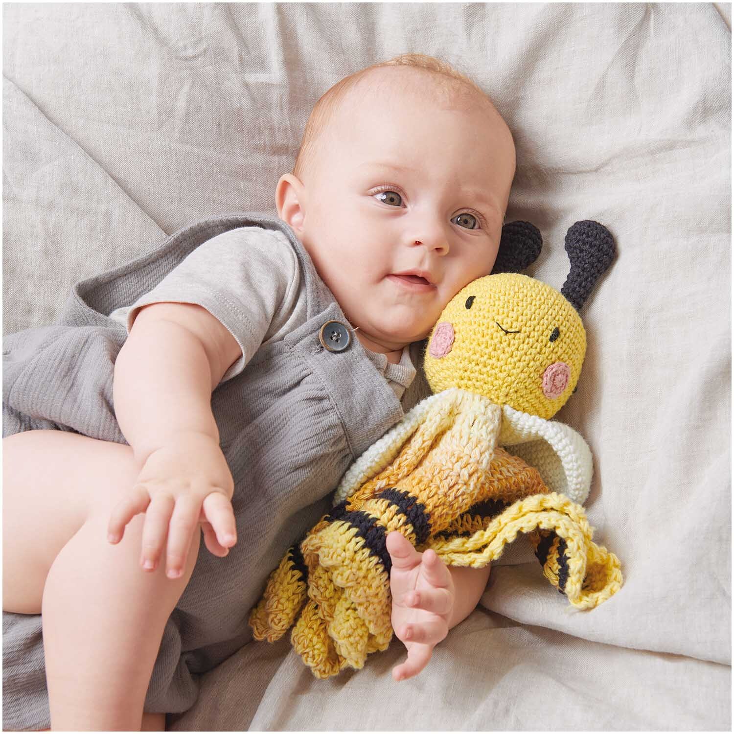 Ricorumi Spin Spin dk: Ein Baby hält ein gelbes, gestricktes Kuscheltier von Rico Design auf einem Bett.