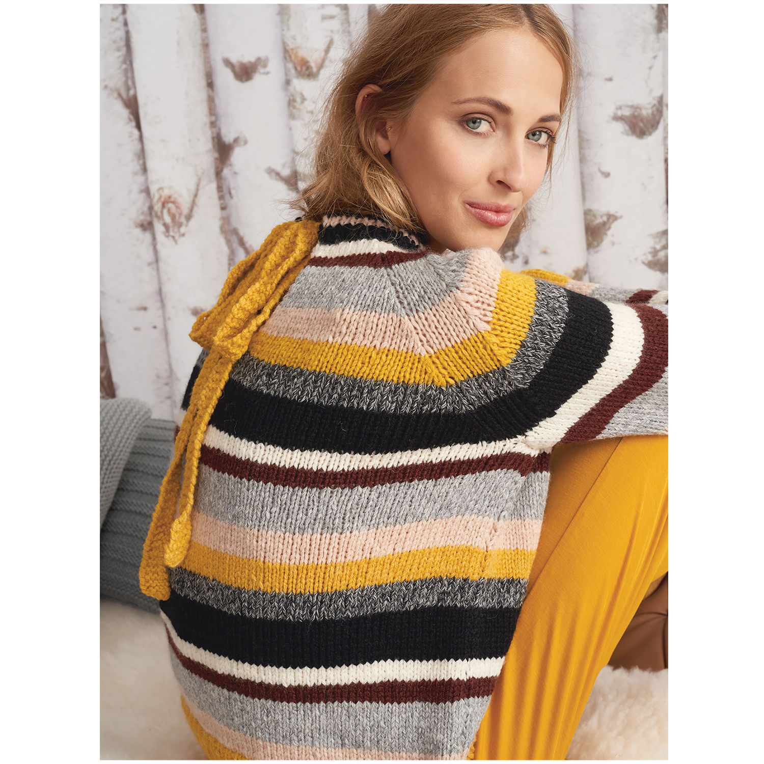 Fashion Alpaca Dream: Frau in gestreiftem Pullover mit Gelb, sitzt vor Birkenwald-Hintergrund.