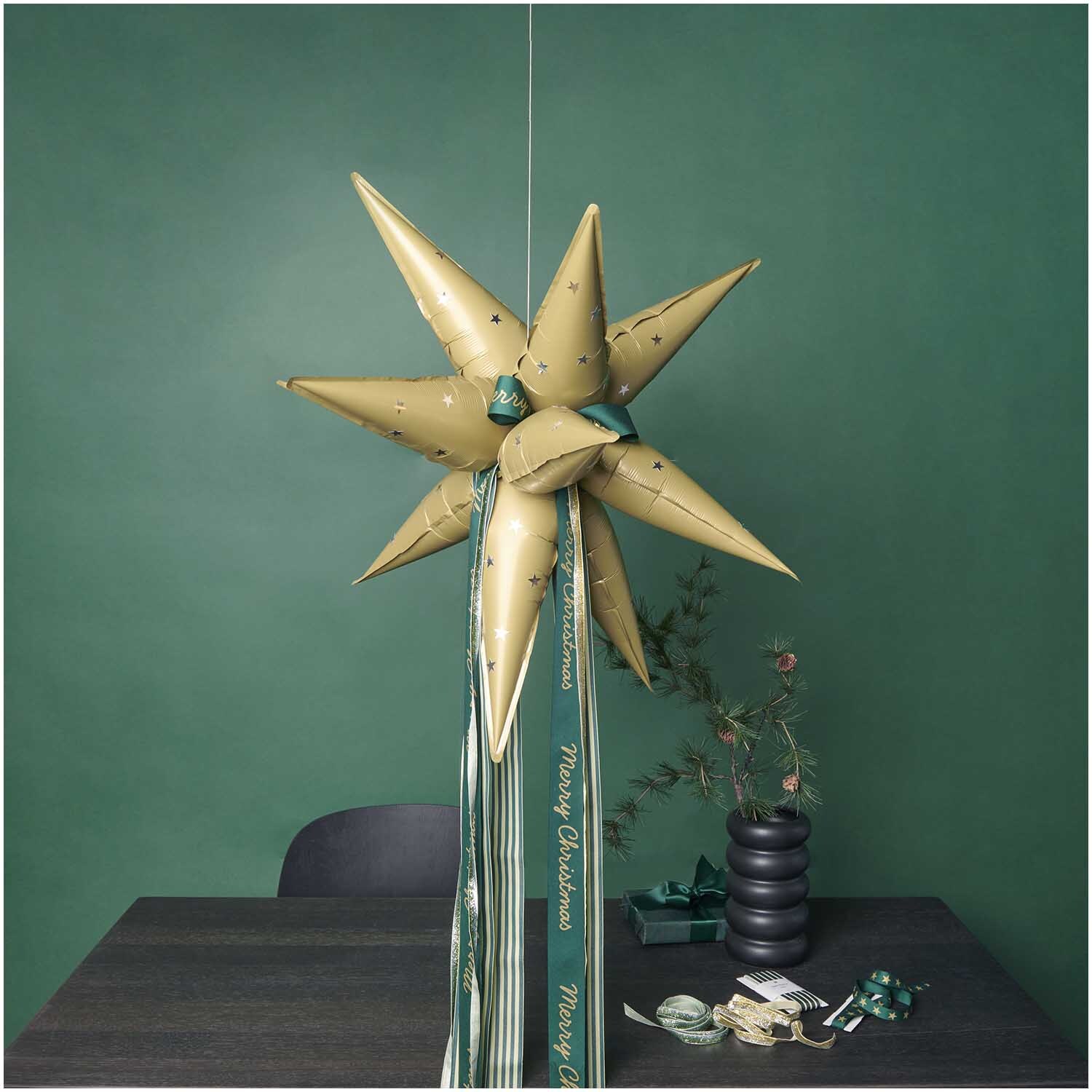 Folienballon 3D-Stern gold von Rico Design schwebt in einem stilvollen Raum mit grüner Wand.