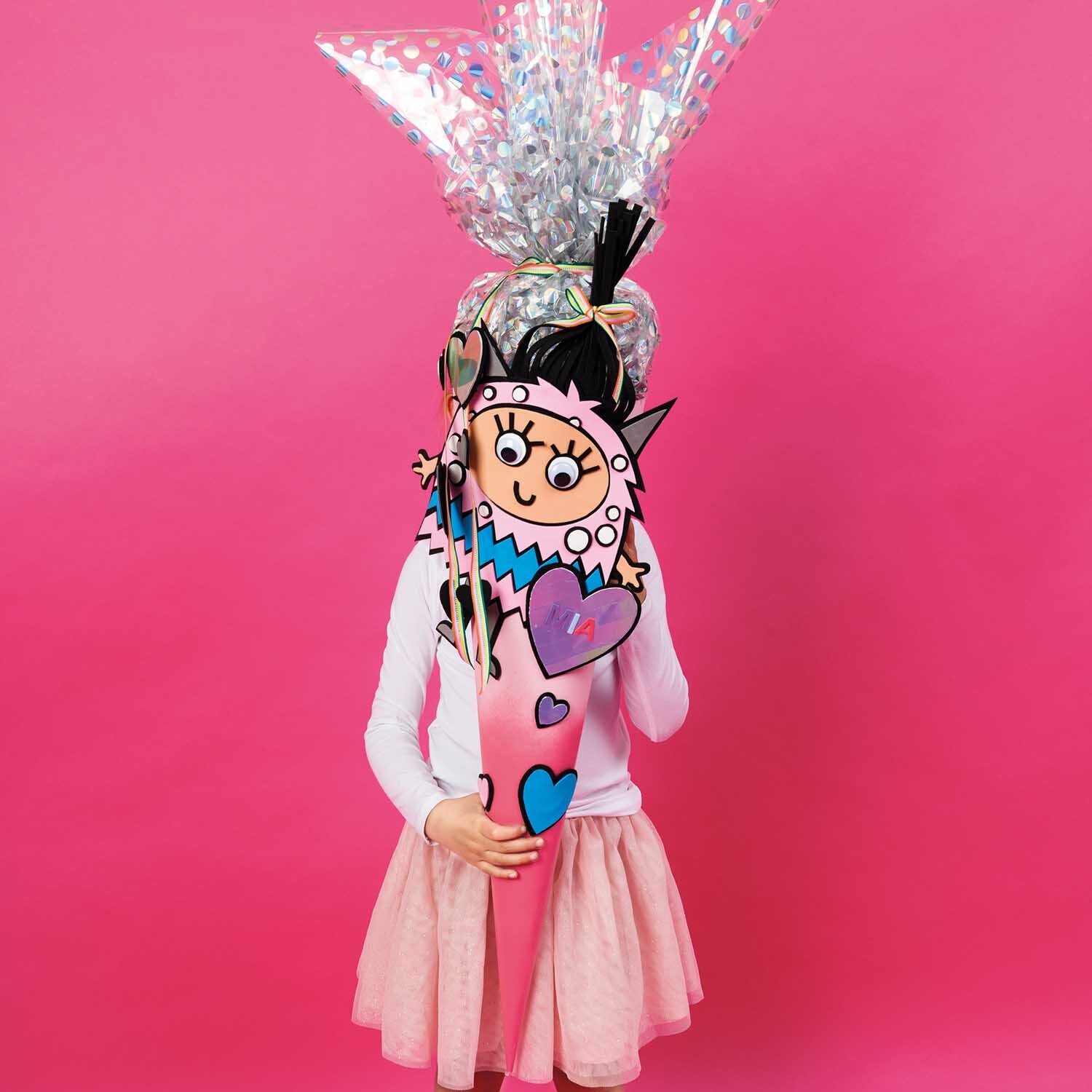 Bastelanleitung Monster-Schultüte für Mädchen: Ein Kind hält eine bunte Schultüte mit Monsterdesign vor einem pinken Hintergrund.