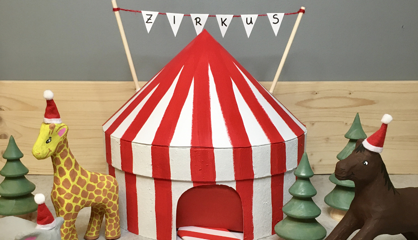 Anleitung Adventskalender Zirkus: Bunter Zirkus mit Tieren und Weihnachtsmützen, umgeben von Tannenbäumen.