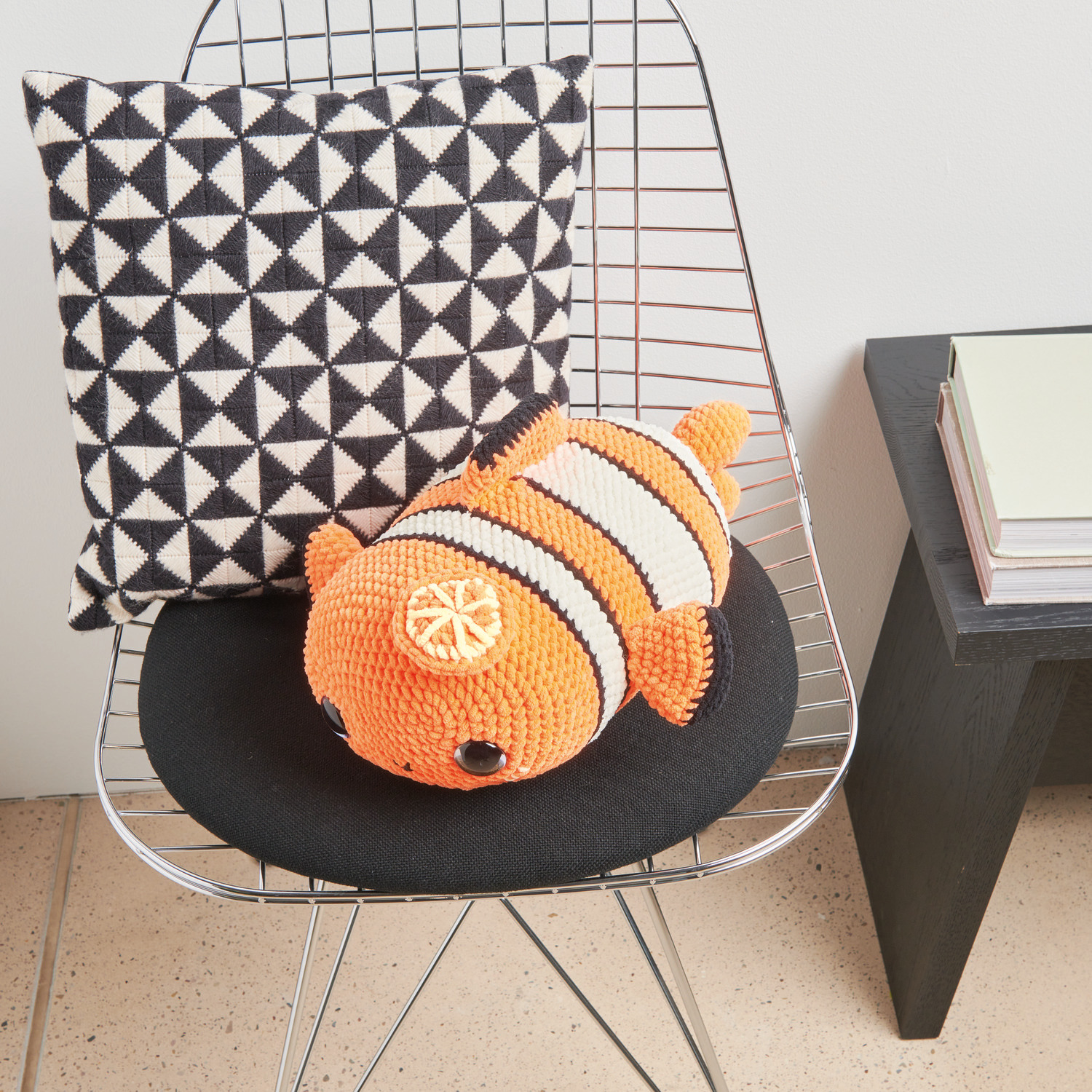 Häkelset Crazy Clown Fish Modell 04: Orange-weißer Fisch aus Chenillove Big Cuties auf Stuhl mit geometrischem Kissen.
