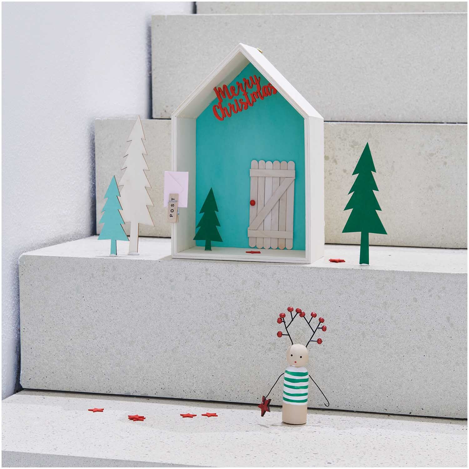 Holzhänger Rentier mit Glitter in einer weihnachtlichen Szene mit Bäumen und einem Haus auf Treppenstufen.