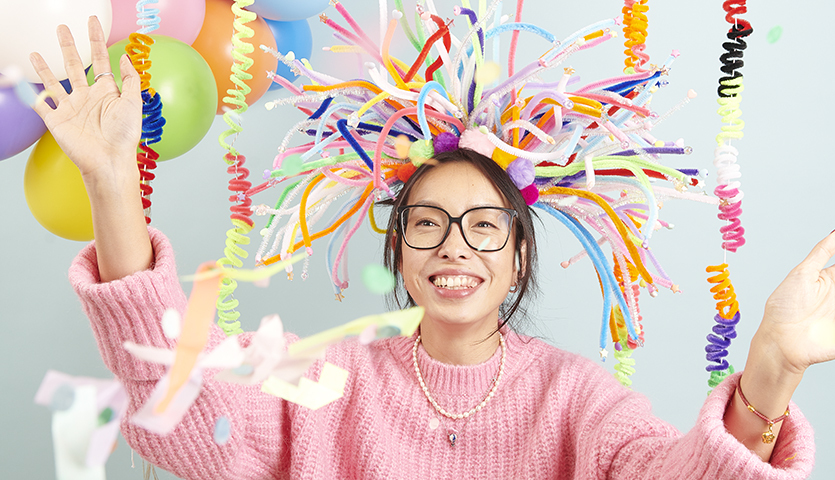 Bastelanleitung Haarreif mit Chenilledraht für Karneval: Frau in rosa Pullover mit buntem Haarreif aus Chenilledraht, umgeben von Luftballons und Girlanden.
