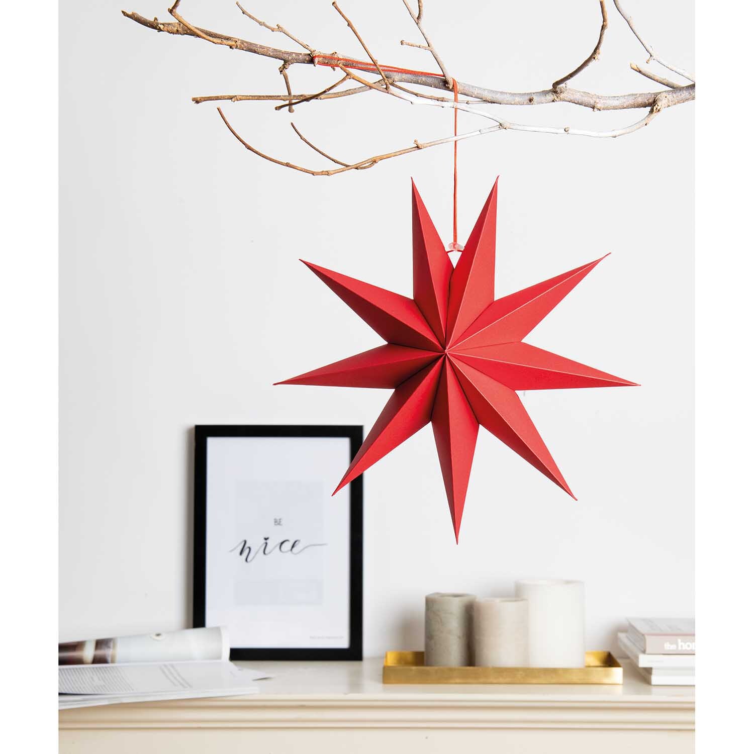 Papierstern auffaltbar 28cm von Rico Design in leuchtendem Rot, hängend an einem Ast über einem Regal mit Kerzen und einem Bild.