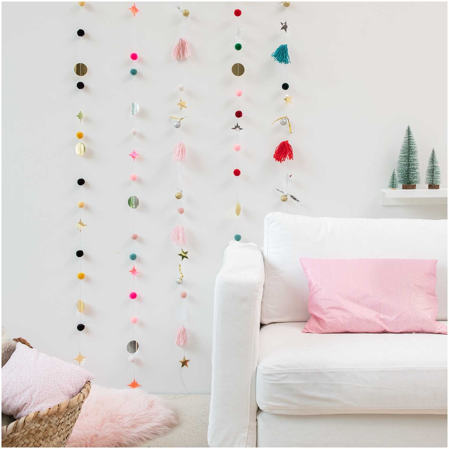 Quasten-Girlande rosa-pastell 2,2m von Rico Design dekoriert eine weiße Wand über einem Sofa.