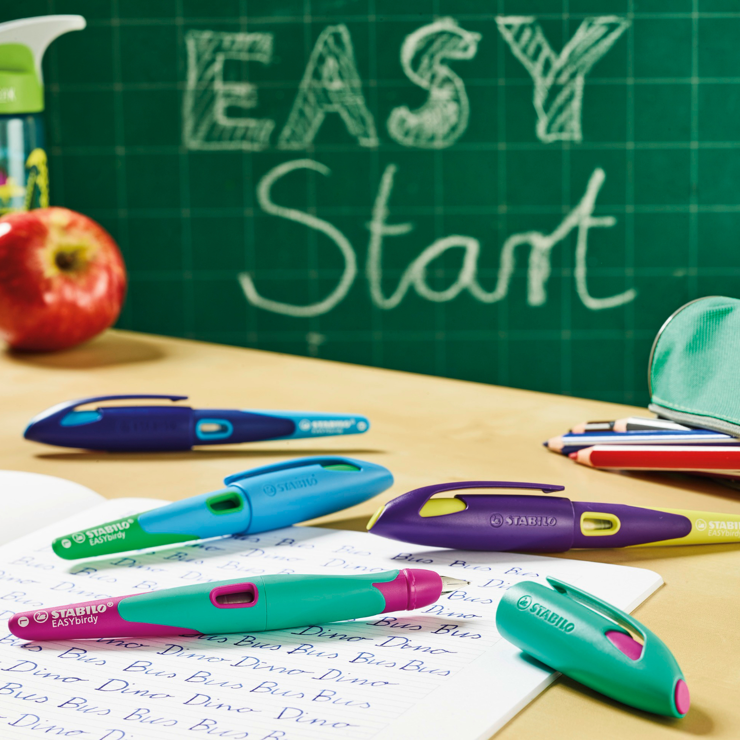 Easybird Patronenfüller für Rechtshänder auf einem Schreibtisch mit Schulmaterialien. Verschiedene Farben, darunter Beere, vor einer grünen Tafel.