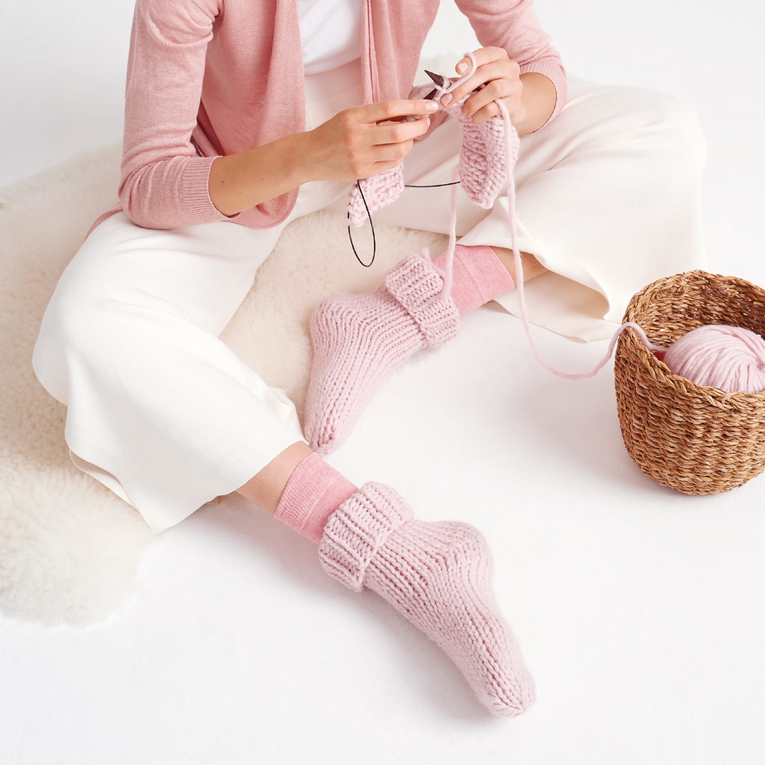 Strickanleitung Socken aus Essentials Super super chunky: Frau strickt rosa Socken, sitzt auf weißem Teppich, neben Korb mit Wolle.