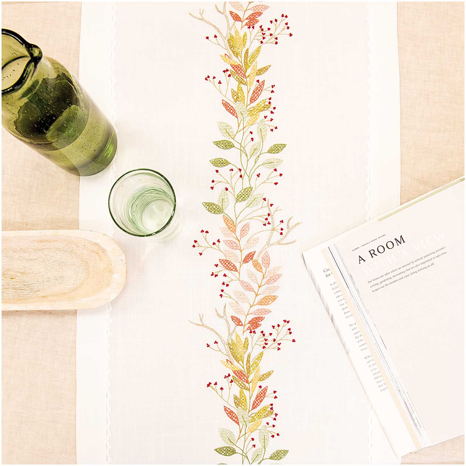 Stickpackung Läufer Herbstkranz 40x150cm zeigt einen Tischläufer mit herbstlichem Blatt- und Beerenmuster, dekoriert mit Glasflaschen und einem Buch.