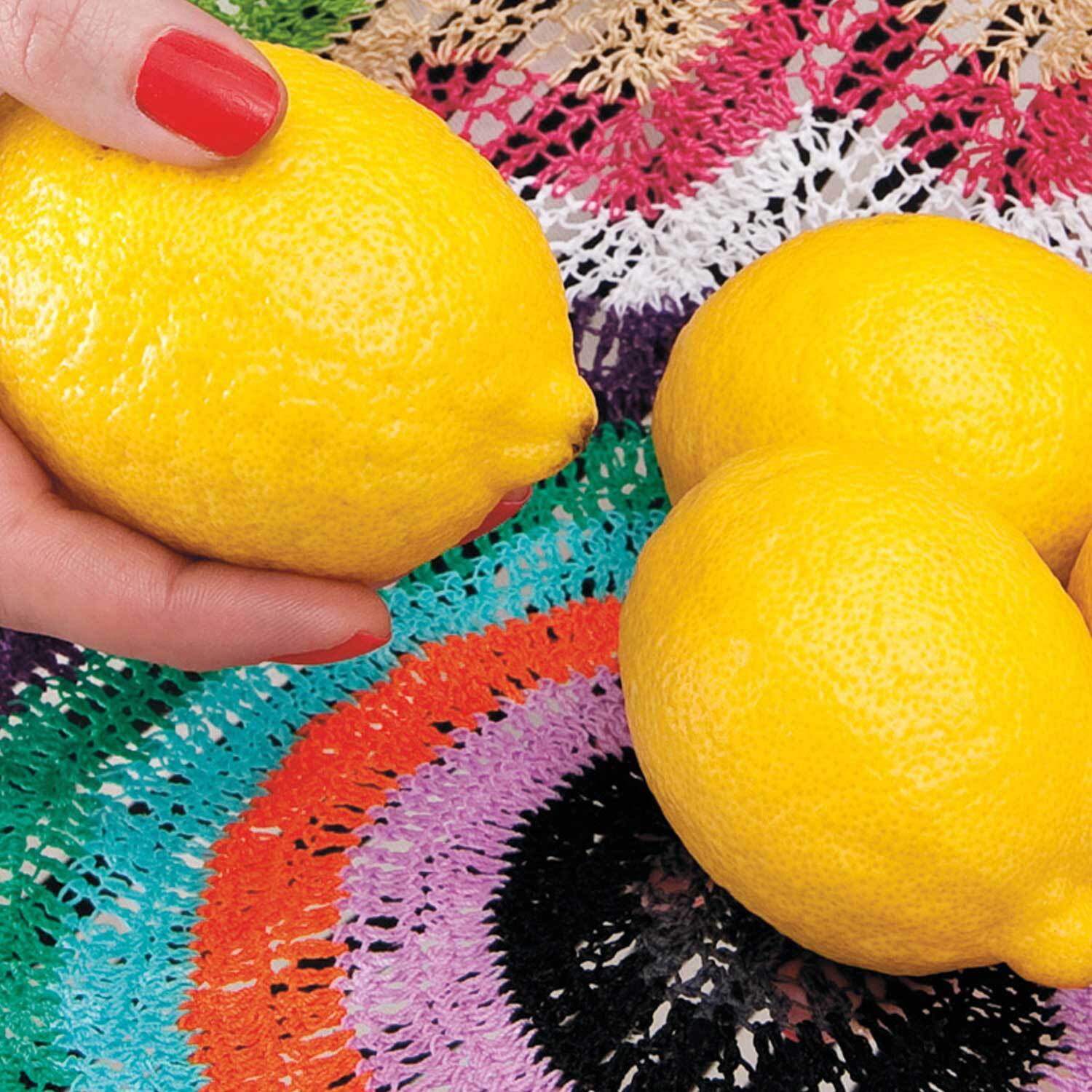 Häkelanleitung Schale aus Häkelgarn: Hand hält Zitrone über bunte, gehäkelte Tischdecke.