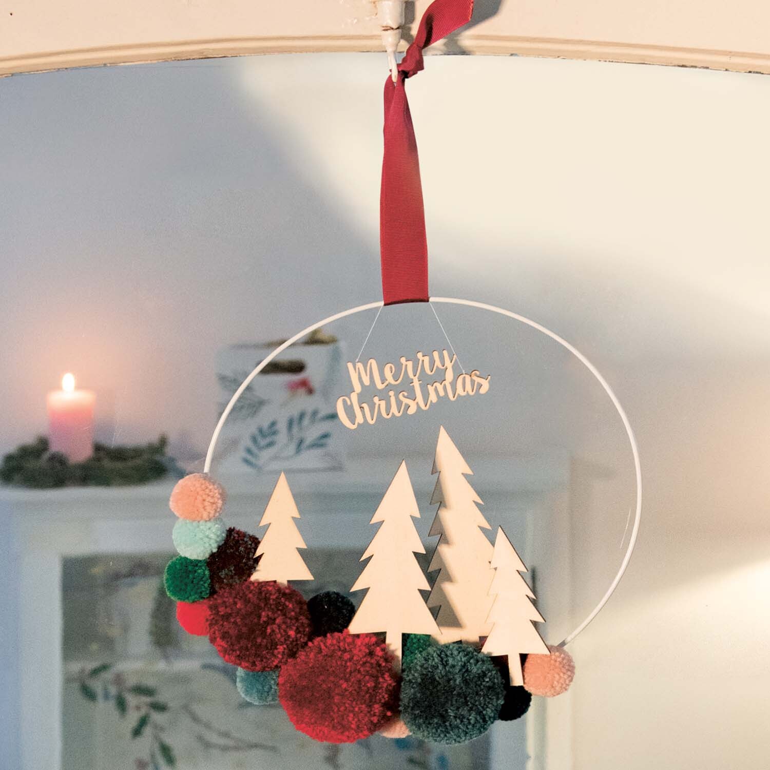 Bastelanleitung weihnachtlicher Pompon-Kranz mit bunten Pompons und Holz-Tannenbäumen, aufgehängt an rotem Band.