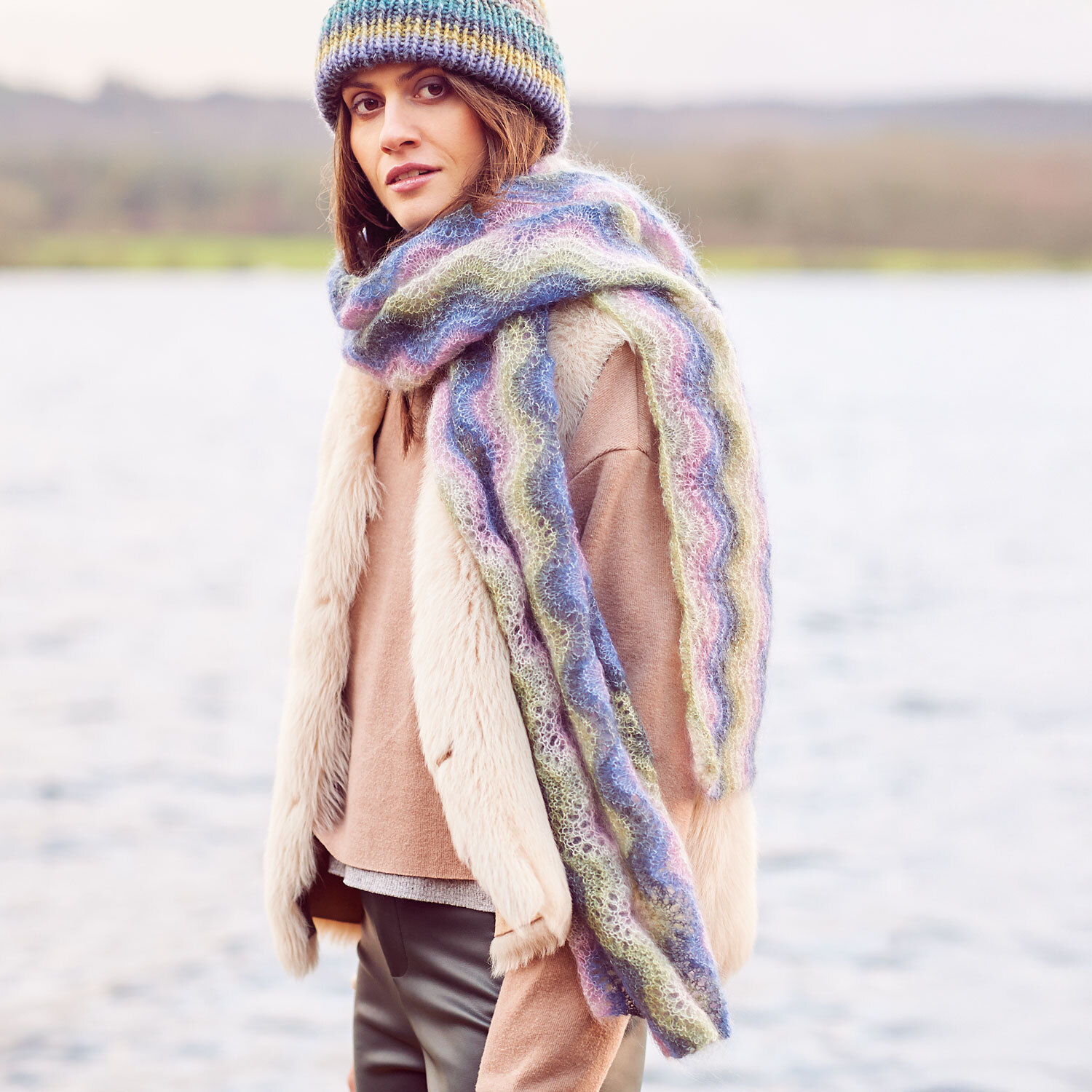 Frau trägt einen farbenfrohen Schal aus Essentials Super Kid Mohair Loves Silk Print. Der Schal zeigt ein wellenförmiges Muster in Pastelltönen. Passend dazu eine Mütze, die das Outfit harmonisch ergänzt.