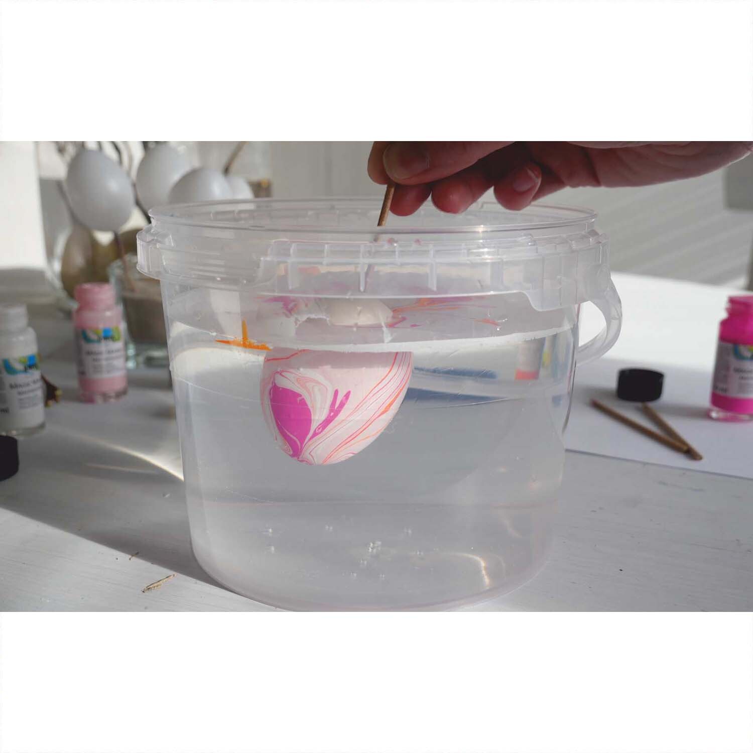 Magic Marble Marmorierfarben Set Love Neon zeigt eine Hand, die ein Ei in einem Wasserbehälter marmoriert.