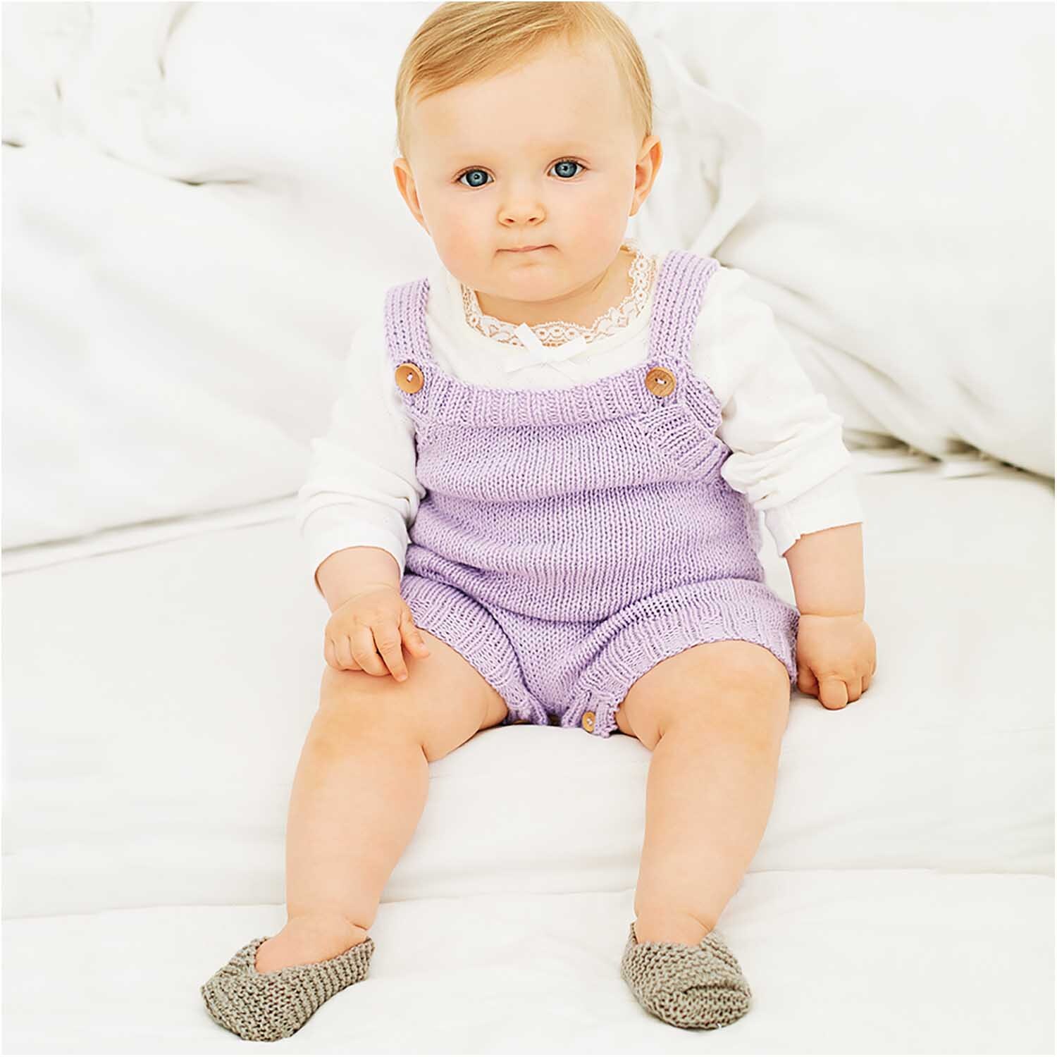 Rico Baby Cotton Soft dk: Ein Baby in lila Strickanzug sitzt auf weißem Hintergrund, trägt graue Schuhe.